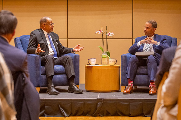 Rev. Dr. Benjamin Chavis and Provost Alec Gallimore sit in blue armchairs on a stage and converse with one another.