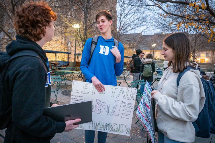 Three students discuss climate change while holding posters and standing on the Bryan Center Plaza.