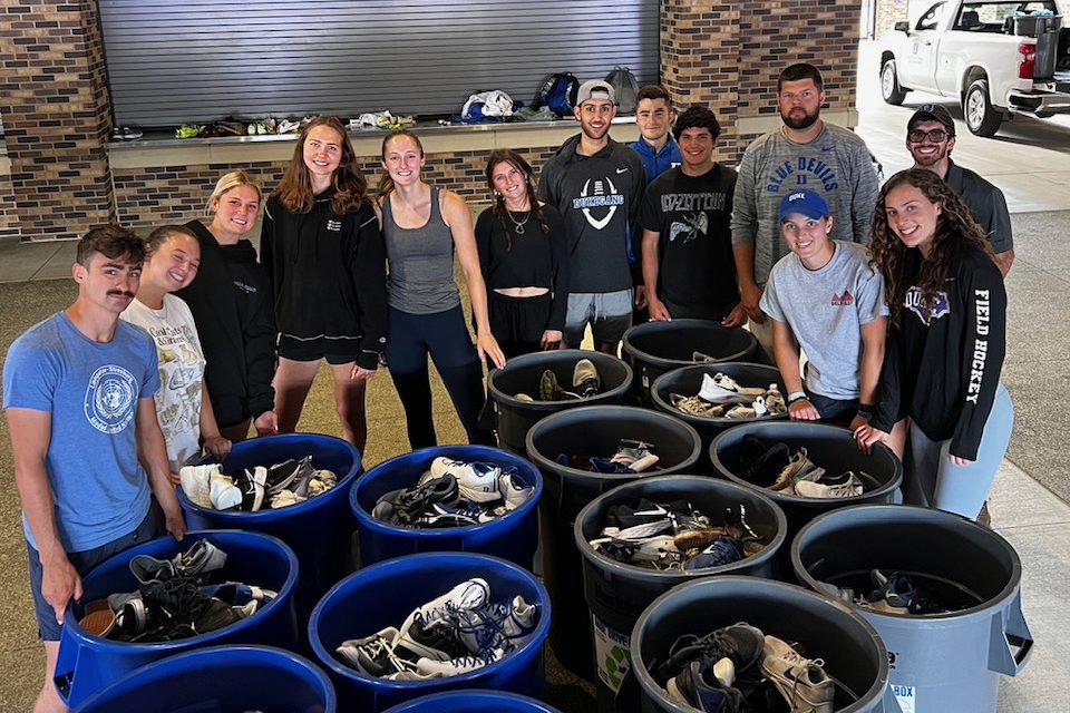 Students standing behind large containers of donated athletics shoes as a part of Duke Sustainable Athletics Group's shoe drive